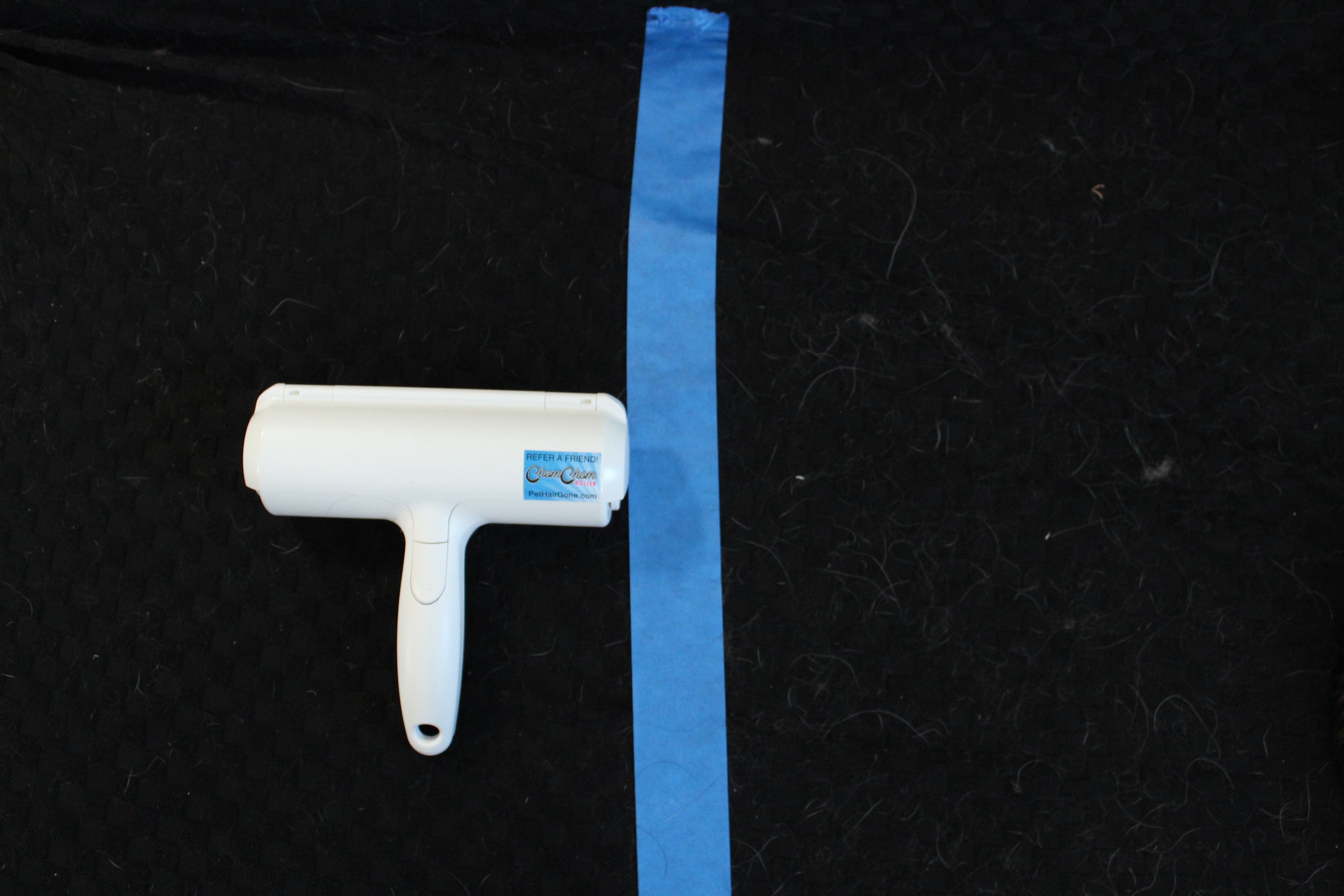 ChomChom pet hair remover roller on a hair-covered black fabric surface, with blue painter's tape marking the test zone