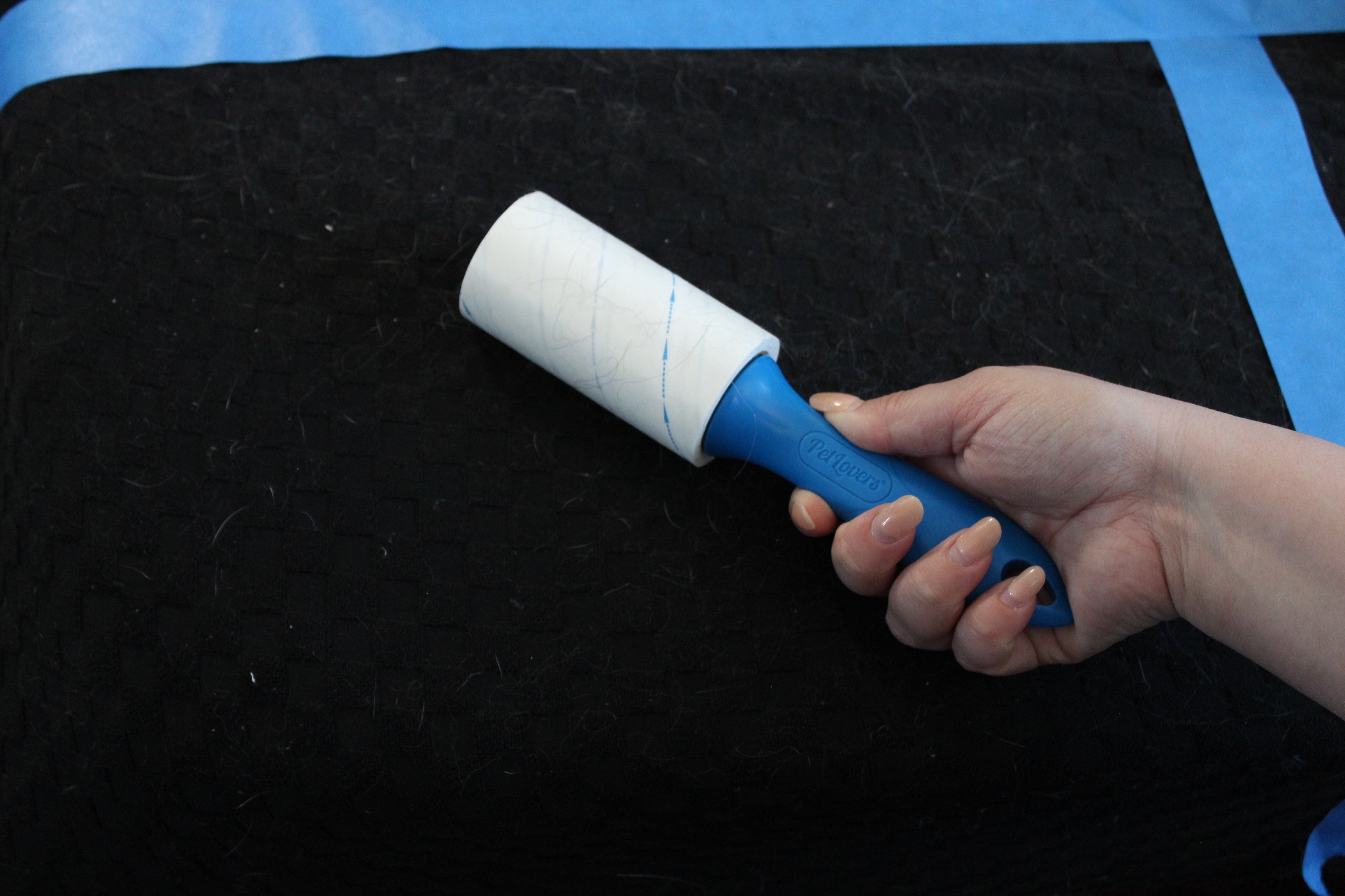 PetLovers Extra Sticky disposable lint roller held over a hair-covered black fabric surface, with cat hair visibly adhered to the sheet after a single pass